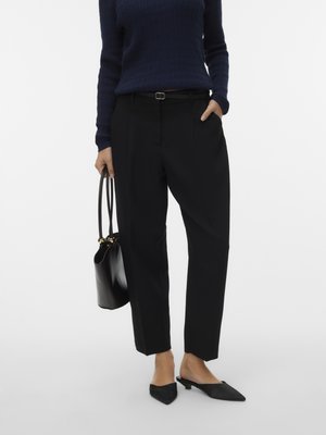 Person wearing black tailored pants, navy sweater, black pointed mule shoes, holding black leather handbag in one hand.