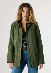 Giacca verde oliva con chiusura a bottoni, due tasche laterali, dettagli marroni e una vestibilità comoda, indossata sopra una camicia a righe.