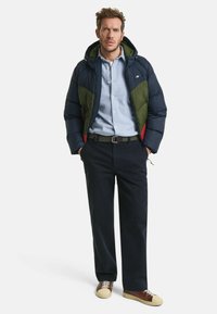 Pufferjack in marineblauw en groen met rode accenten, ritssluiting en verstelbare capuchon. Gecombineerd met een lichtblauw shirt en donkere broek.