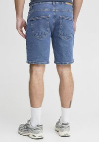 Shorts de denim en azul medio, con un corte clásico y dobladillo deshilachado. Dos bolsillos traseros y un detalle de trabilla para cinturón. Combinados con zapatillas gris.