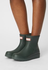 Botas de tobillo de goma verdes con un acabado mate, que presentan un logo en rojo y blanco, puntera redonda y suela texturizada para mayor agarre.