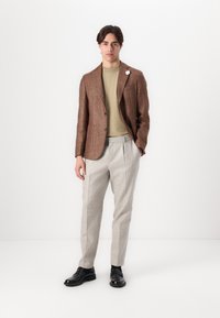 Hnedý textúrovaný blazer cez zelený top, v kombinácii so svetlošedými kockovanými nohavicami a čiernymi topánkami. Výrazný špendlík na klopu blazeru.