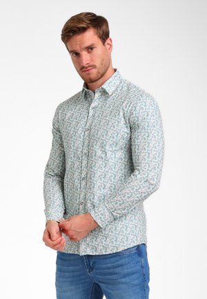 Man fastening cuffs of light-colored button-up shirt with pastel geometric pattern, wearing blue jeans, against plain white background.