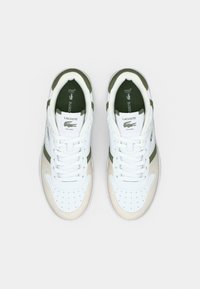 Ζευγάρι λευκά sneakers Lacoste με μπεζ και πράσινες λεπτομέρειες, διάτρητο μπροστινό μέρος και γλώσσα με λογότυπο, σε προοπτική από πάνω σε απλό φόντο.