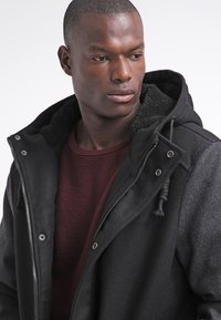 Schwarze Kapuzenjacke mit Reißverschluss, ausgestattet mit einem weichen, strukturierten Innenfutter und Kordelzügen. Getragen über einem burgunderfarbenen Pullover.