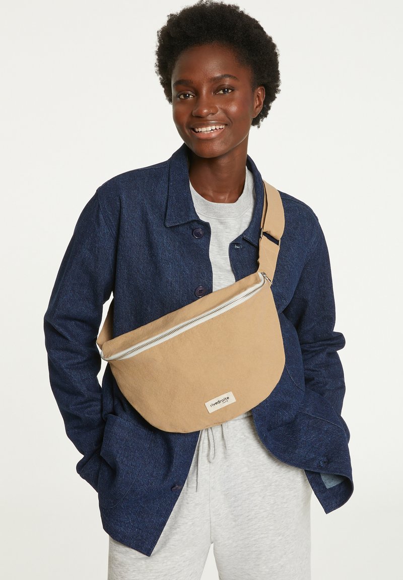 Sac en toile beige à bandoulière avec une fermeture éclair argentée, porté sur une veste bleue à boutons, présentant une étiquette à l'avant et un design simple.