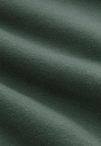 Tela de punto verde con una textura suave, presenta un fin ribeteado y un brillo sutil, exhibida en pliegues para mostrar profundidad y flexibilidad.