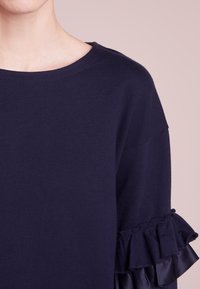 Suéter de punto azul marino con un escote redondo y un detalle de volantes en las mangas, hecho de una tela suave. Textura suave con un detalle sutil.