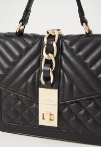 Borsa nera trapuntata con hardware dorato, un accento in catena e una chiusura rettangolare impresso con il nome del marchio "ALDO". Texture liscia.