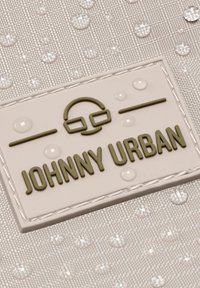 Fehér textúrájú anyag, amelyen emelt zöld betűk formájában a 'Johnny Urban' logó látható, fényes vízcseppekhez hasonló foltokkal körülvéve.