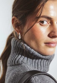 Knit gray turtleneck sweater with ribbed texture, paired with a teardrop-shaped silver earring. Smooth skin and pulled-back hair are visible.