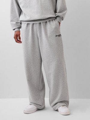 Person trägt lockere graue Sweatpants mit dem Schriftzug "gap·awake new york" in der Nähe der Tasche, kombiniert mit weißen Sneakers und passendem grauen Sweatshirt.