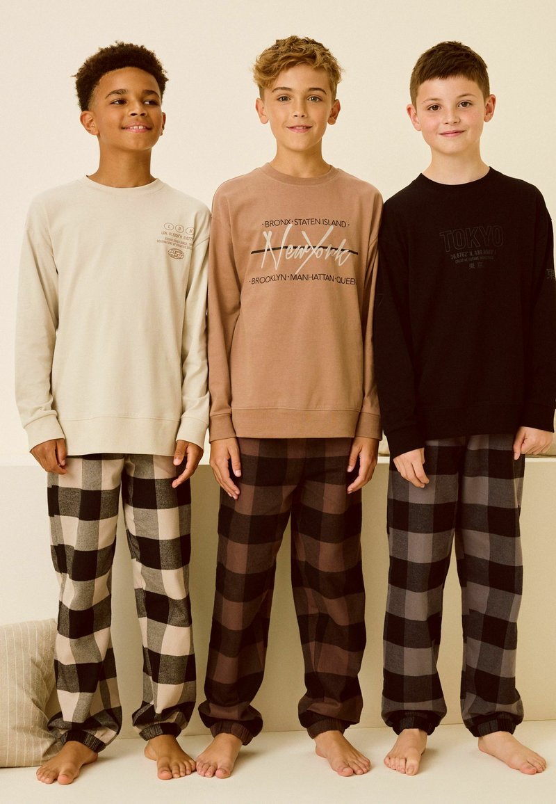 Trois garçons portent des chemises à manches longues et des pyjamas en flanelle. Les chemises sont beige, marron clair et noir ; les pantalons présentent différents motifs à carreaux dans des couleurs sombres.