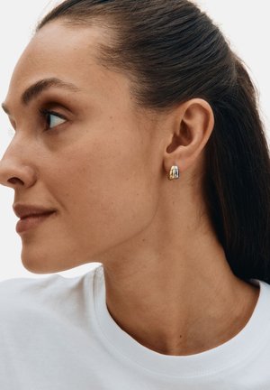 Gold and silver hoop earrings on a model, featuring a smooth, curved design with polished surfaces. Model has long hair and wears a white shirt.