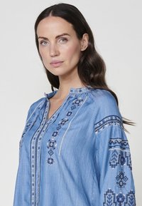 Blå chambrayblus med intrikata blå och silverbroderier längs knäppningen och ärmarna. Har en mandarinkrage och en avslappnad passform.