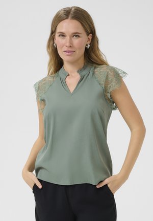 Blusa verde con maniche corte in pizzo, un colletto arricciato discreto e una vestibilità comoda. Tessuto liscio con una leggera lucentezza.