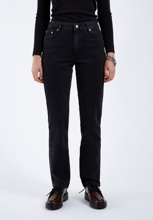 Person iført sorte jeans med højt talje og lige ben, sort langærmet top, brune snørestøvler og sølvkæde armbånd på venstre håndled.