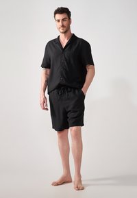 Svart lin skjorte med korte ermer, knappeforskyvning og brystlomme, kombinert med matchende svarte shorts med snøring, vist på en stående figur.