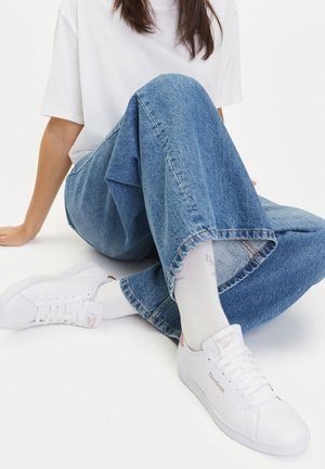 Białe trampki Reebok z teksturowaną skórą, detalami sznurowania i wytłoczonym logo, w zestawieniu z jasnoniebieskimi szerokimi spodniami dżinsowymi i białymi skarpetami.