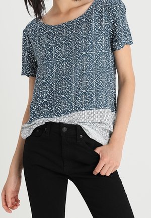 Woman wearing a blue patterned short-sleeve top over a white patterned undershirt and black pants, hand in pocket.
