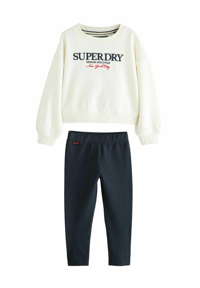 Kremeni dukserica s "SUPERDRY" logom, u kombinaciji s tamnoplave tajice. Mekani pamučni materijal, opušten kroj, rebrasti rukavi i pojas.