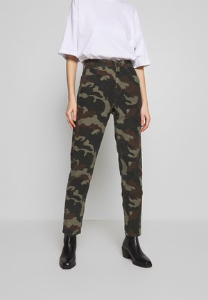 Person iført en løs hvid T-shirt, camo-mønstrede bukser og sorte ankelstøvler, stående mod en ensfarvet lysegrå baggrund.