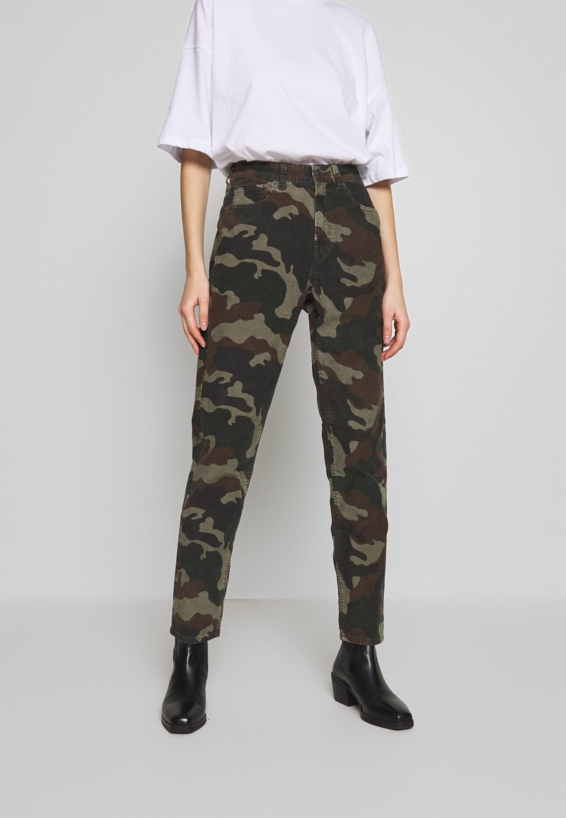 Personne portant un T-shirt blanc ample, un pantalon à motif camouflage et des bottines noires, debout devant un fond gris clair uni.