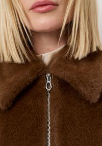 Bruine faux bont jas met een rits aan de voorkant en een kraag. Kenmerkend zijn de zilveren rits en de zachte textuur. Dichtbijzicht, met een nadruk op de details van de stof.