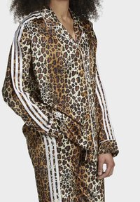 Pajamaset i leopardmönster i glänsande satin, med kontrasterande vita ränder längs ärmarna. Knappar och fickor är framträdande designdetaljer.