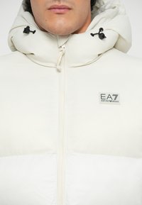 White padded jacket with a high collar, front zipper, and adjustable drawstrings. Features a logo patch on the left side.