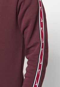 Maroon sweatshirt with a ribbed texture, featuring a white and red stripe along the sleeve with repeated Nike logos. Simple, fitted design.