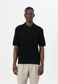 Μαύρη πλεκτή πόλο μπλούζα με κοντά μανίκια, γιακά και πλέγμα με τρία κουμπιά. Φοριέται με ελαφρώς μπεζπλισέ παντελόνι.