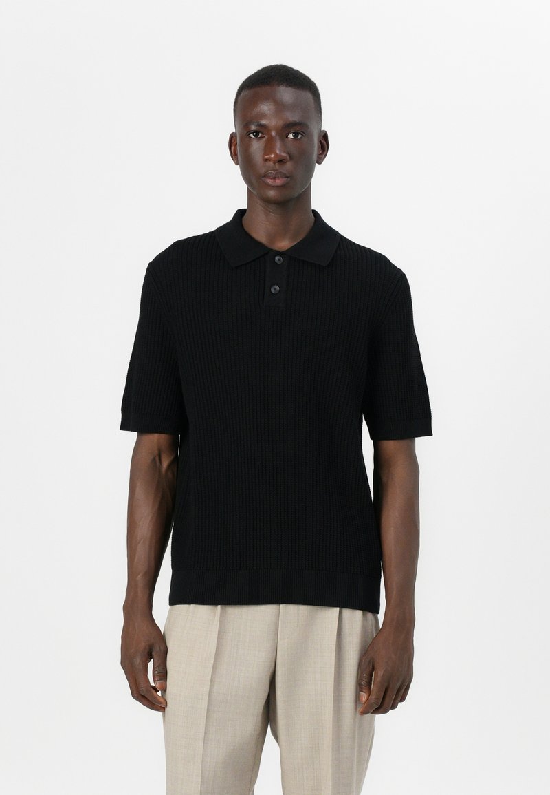 Μαύρη πλεκτή πόλο μπλούζα με κοντά μανίκια, γιακά και πλέγμα με τρία κουμπιά. Φοριέται με ελαφρώς μπεζπλισέ παντελόνι.