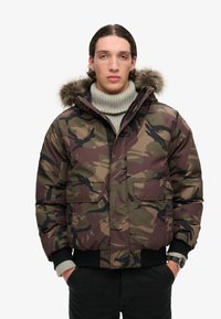 Chaqueta acolchada de camuflaje con capucha forrada de piel extraíble, cierre de cremallera en la parte delantera, dos bolsillos en el pecho, puños acanalados y tejido texturizado en tonos terrosos.