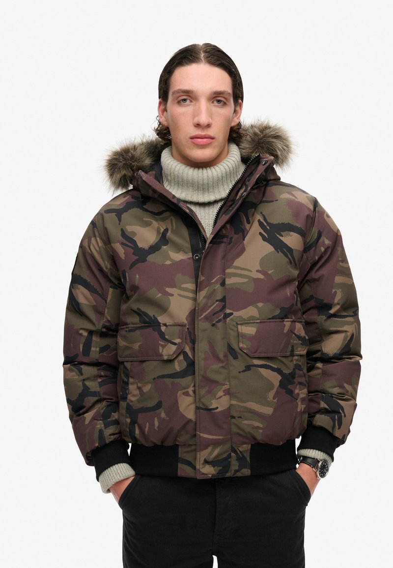 Chaqueta acolchada de camuflaje con capucha forrada de piel extraíble, cierre de cremallera en la parte delantera, dos bolsillos en el pecho, puños acanalados y tejido texturizado en tonos terrosos.