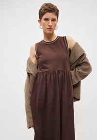 Καφέ φόρεμα με εφαρμοστή μέση και φουσκωτή φούστα, με στρογγυλή λαιμόκοψη και λευκό ραφές. Συνδυάζεται με απαλό, μπεζ ζακέτα.