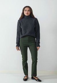 Dunkelgrauer gestrickter Rollkragenpullover mit gerippten Bündchen, kombiniert mit eng anliegenden olivgrünen Hosen und schwarzen Ballerinas, vor einem neutralen Hintergrund stehend.