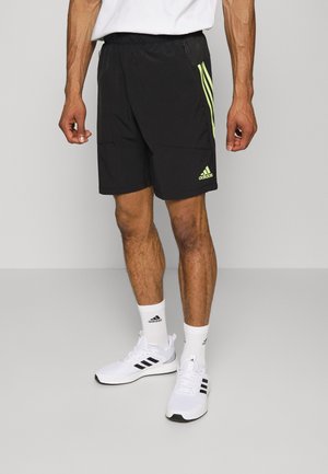 Zwarte sportshorts met neon groene strepen, elastische tailleband en zijzakken. Draagt witte sokken en schoenen met drie zwarte strepen.