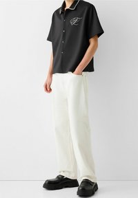 Czarna krótka koszula na guziki z białym obszyciem kołnierzyka i haftowanym logo, połączona z białymi szerokimi spodniami i czarnymi masywnymi butami.