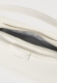 Λευκή τσάντα Calvin Klein με λείο, υφασμένο εξωτερικό, κορυφαίο κλείσιμο με φερμουάρ και γκρι εσωτερικό. Διαθέτει λογότυπο σε δερμάτινη λεπτομέρεια.