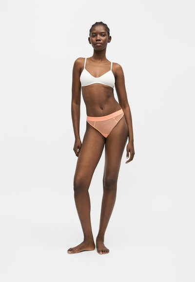 Γυναίκα στέκεται ξυπόλητη φορώντας λευκό bralette και εσώρουχο μπικίνι από ροδακινί δίχτυ σε απλό ανοιχτό γκρι φόντο.