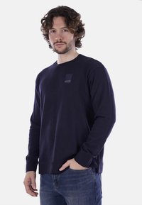 Maglione lavorato a maglia blu navy con polsini e orlo a coste, caratterizzato da una piccola etichetta testurizzata sul petto. Texture liscia, design relax.