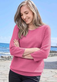 Frau mit langem grauem Haar, die einen strukturierten rosa Pullover und schwarze Hosen trägt, steht mit verschränkten Armen nahe einem Steinweg am Meer.