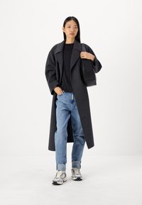 Cappotto lungo in lana grigio antracite con revers oversize, t-shirt nera, jeans leggermente strappati di colore blu chiaro e sneakers argento/nero. Borsa a tracolla nera.
