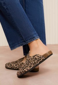 Dierenprint klompen met een beige en zwart luipaardpatroon, kurken zool, verstelbare gesp en een rond toelopend ontwerp.