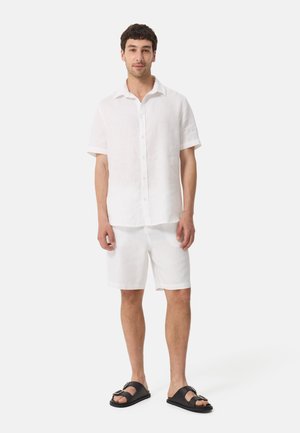 Witte linnen overhemd met korte mouwen en bijpassende shorts met een ontspannen pasvorm, gecombineerd met zwarte slippers, tegen een eenvoudige achtergrond.