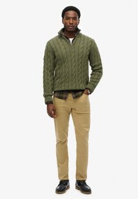 Pull en maille torsadée verte avec col zippé, superposé à une chemise à carreaux, associé à un pantalon beige et des bottes noires. Design simple et texturé.
