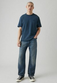 T-shirt in cotone blu con scollatura rotonda, abbinata a jeans in denim azzurro chiaro. L'outfit presenta sneakers a motivo bianco e nero.