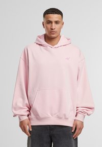 Haut à capuche oversized rose clair en tissu doux, doté d'une poche avant et d'une capuche avec cordon de serrage. Petit logo en rose contrasté sur la poitrine.
