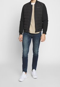 Czarna puchowa kurtka bomberka o gładkim wykończeniu, z zamkiem błyskawicznym i ściągaczami przy mankietach; noszona na beżowej koszuli i zestawiona z ciemnymi dżinsami oraz białymi trampkami.